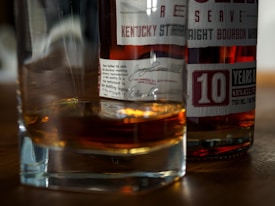 A close-up view of a glass of whiskey alongside a bottle of bourbon with visible labels detailing its contents and age. The label indicates '10 years old' and mentions the alcohol content. The scene is set on a wooden surface, and the whiskey in the glass casts a warm, amber reflection.