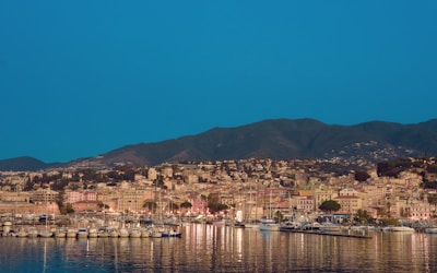 A coastal cityscape with a marina filled with numerous docked sailboats. The city is nestled against a backdrop of rolling hills, with a mix of pastel-colored buildings and greenery. The water in the foreground reflects the boats and city lights.