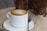A frothy cappuccino served in a white cup placed on a matching saucer with a metal spoon beside it. The setting includes a sandy surface, adding an outdoor, beach-like vibe with the base of a tree visible in the background.