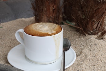 A frothy cappuccino served in a white cup placed on a matching saucer with a metal spoon beside it. The setting includes a sandy surface, adding an outdoor, beach-like vibe with the base of a tree visible in the background.