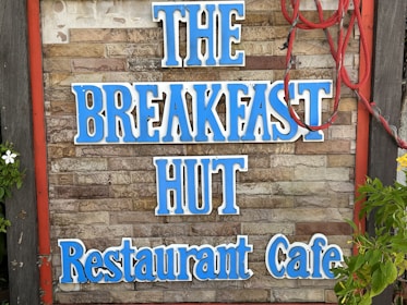 A rustic sign on a brick wall displays the words 'THE BREAKFAST HUT Restaurant Cafe' in bold blue letters. Red rope decorates the right side of the sign, and there are green leaves and white flowers visible on the left.