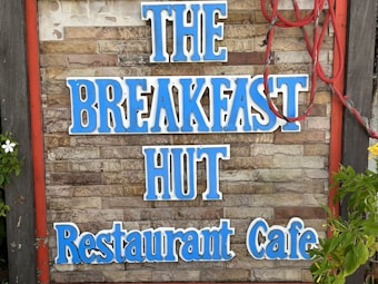 A rustic sign on a brick wall displays the words 'THE BREAKFAST HUT Restaurant Cafe' in bold blue letters. Red rope decorates the right side of the sign, and there are green leaves and white flowers visible on the left.