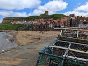 A coastal town features a sandy beach in the foreground where people are relaxing. Colorful residential buildings with red roofs line the beachfront. A grassy hill rises behind the houses, topped with a historic church or abbey. There are lobster traps in the foreground, suggesting a fishing community.