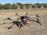 A large drone with multiple propellers is positioned on a patch of dry grass. It appears to be well-equipped, possibly for agricultural or industrial purposes, given its size and design. Behind the drone, there is a glimpse of a road and some solar panels, with a background of shrubs and trees under a clear blue sky.
