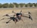 A large drone with multiple propellers is positioned on a patch of dry grass. It appears to be well-equipped, possibly for agricultural or industrial purposes, given its size and design. Behind the drone, there is a glimpse of a road and some solar panels, with a background of shrubs and trees under a clear blue sky.