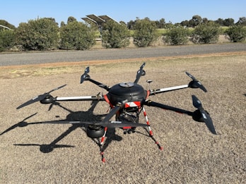 A large drone with multiple propellers is positioned on a patch of dry grass. It appears to be well-equipped, possibly for agricultural or industrial purposes, given its size and design. Behind the drone, there is a glimpse of a road and some solar panels, with a background of shrubs and trees under a clear blue sky.