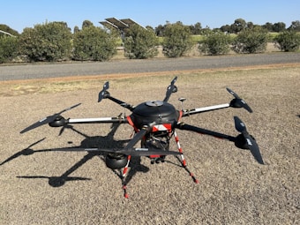 A large drone with multiple propellers is positioned on a patch of dry grass. It appears to be well-equipped, possibly for agricultural or industrial purposes, given its size and design. Behind the drone, there is a glimpse of a road and some solar panels, with a background of shrubs and trees under a clear blue sky.