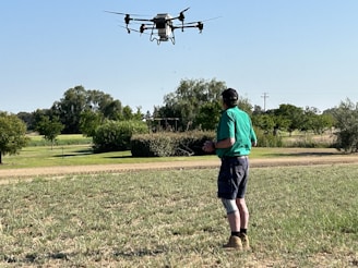 A person standing on a grassy field is operating a drone with a remote controller. The drone is hovering in the air, and trees and shrubs are visible in the background under a clear blue sky.
