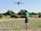 A woman standing in a lush green field holding a drone controller, with crops in the background.