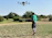 A farmer in a wide green field operating a DJI Agras drone with a remote control.