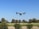 A drone hovers in the sky above a landscape of green fields and trees under a clear blue sky.