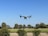 A drone hovers in the sky above a landscape of green fields and trees under a clear blue sky.