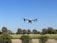 A drone hovers in the sky above a landscape of green fields and trees under a clear blue sky.