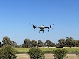 A drone hovers in the sky above a landscape of green fields and trees under a clear blue sky.