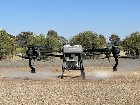 A large drone is hovering above a dry grassy field, releasing a mist or spray. The background features green bushes and trees under a clear blue sky.