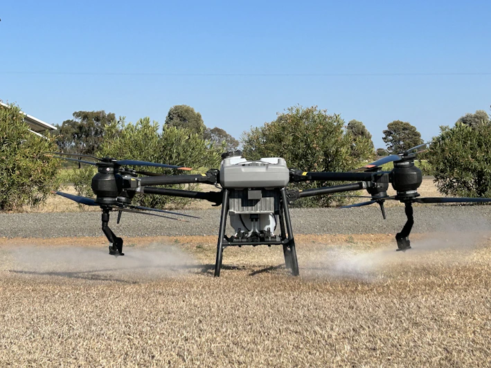 A sleek agricultural drone hovering low over a lush green field, releasing a fine mist of spray with a backdrop of clear blue skies.