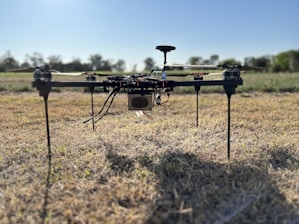 A quadcopter drone is resting on a field of dry grass. The drone features four rotors and appears to have various components and wires exposed. The background shows a clear sky and a blurred treeline, indicating an open outdoor setting.