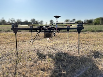 A quadcopter drone is resting on a field of dry grass. The drone features four rotors and appears to have various components and wires exposed. The background shows a clear sky and a blurred treeline, indicating an open outdoor setting.