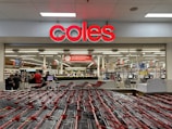 A supermarket entrance with a prominent red sign displaying 'coles' above the entryway. Numerous shopping carts are lined up in front of the store. Inside, various aisles can be seen, stocked with products under bright fluorescent lighting. Several people are visible, engaging in shopping activities.