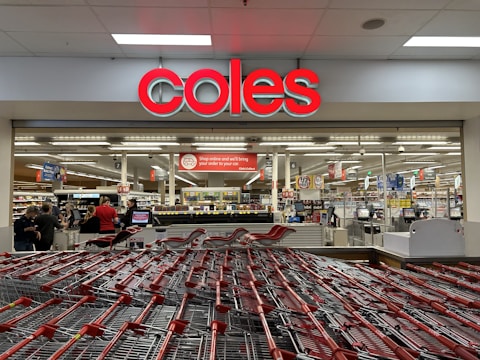 A supermarket entrance with a prominent red sign displaying 'coles' above the entryway. Numerous shopping carts are lined up in front of the store. Inside, various aisles can be seen, stocked with products under bright fluorescent lighting. Several people are visible, engaging in shopping activities.