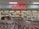 A supermarket entrance with a prominent red sign displaying 'coles' above the entryway. Numerous shopping carts are lined up in front of the store. Inside, various aisles can be seen, stocked with products under bright fluorescent lighting. Several people are visible, engaging in shopping activities.