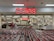 A supermarket entrance with a prominent red sign displaying 'coles' above the entryway. Numerous shopping carts are lined up in front of the store. Inside, various aisles can be seen, stocked with products under bright fluorescent lighting. Several people are visible, engaging in shopping activities.