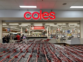 A supermarket entrance with a prominent red sign displaying 'coles' above the entryway. Numerous shopping carts are lined up in front of the store. Inside, various aisles can be seen, stocked with products under bright fluorescent lighting. Several people are visible, engaging in shopping activities.