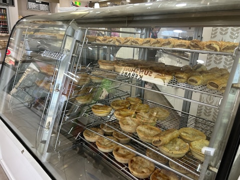 A display case filled with various baked goods including pies and rolls. The shelves are neatly organized with different types of pastries, some topped with seeds and others with a golden brown crust. The background features a partially visible chalkboard menu and decor with leaf patterns.