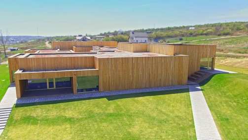 A modern, minimalist wooden building with a geometric design stands amidst a grassy landscape. The structure features flat roofs, large windows, and pathways leading to the entrances. The surrounding area includes grassy fields and sparse residential buildings in the distance.