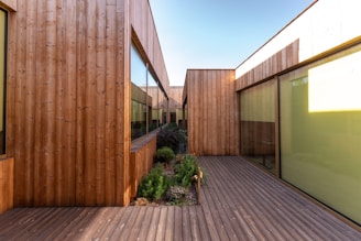 A modern architectural structure features walls and walkways made of natural wood. Large glass windows are incorporated into the design, providing a sleek and contemporary look. The space between the structures includes a small garden with various green plants, adding a touch of nature. The clear sky above suggests clarity and openness.