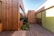 A modern architectural structure features walls and walkways made of natural wood. Large glass windows are incorporated into the design, providing a sleek and contemporary look. The space between the structures includes a small garden with various green plants, adding a touch of nature. The clear sky above suggests clarity and openness.