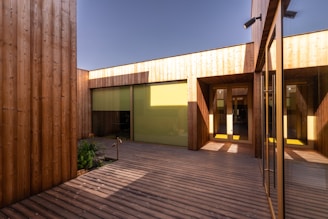 A modern courtyard with wooden walls and a deck, featuring large yellow-tinted windows and glass doors. The wooden panels add a natural and warm aesthetic, while the geometric design and clean lines give a contemporary feel. Sunlight casts shadows across the deck, enhancing the structure's features.