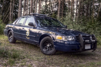 A dark blue state trooper patrol car is parked in a wooded area, surrounded by tall trees. The vehicle has a visible badge on the side and is equipped with a bull bar and roof lights.