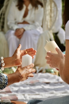 Several people are gathered, sitting on a blanket outdoors. Two individuals are holding white candles, likely participating in a gentle ritual or ceremony. A person wearing white clothing is in the background, suggesting a relaxed atmosphere.