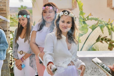 Three women are outdoors, wearing flower crowns and light-colored clothing. The woman in the forefront is holding a smoking bundle, possibly sage, creating a mist around her. They appear relaxed and are participating in a ritual or celebration.
