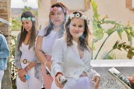 Three women are outdoors, wearing flower crowns and light-colored clothing. The woman in the forefront is holding a smoking bundle, possibly sage, creating a mist around her. They appear relaxed and are participating in a ritual or celebration.
