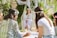 A group of women wearing floral headbands are gathered outdoors, surrounded by lush greenery. One woman sits on a hanging chair in white attire while others, seated on a blanket, engage with candles.