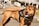 A brown dog wearing a black harness labeled 'Police' stands outdoors on a paved surface. Its mouth is open with its tongue out, and it appears alert. In the background, there are industrial buildings, greenery, and a parked truck.