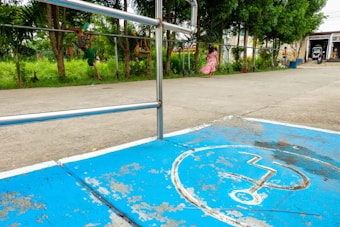 A faded blue handicapped parking symbol is painted on the concrete ground. A metal rail separates a paved area from a grassy section with trees. In the background, a few people, including a child wearing a pink dress, are walking near the trees.