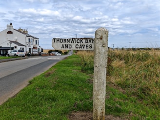 A countryside road with a signpost marking the direction to Thornwick Bay and Caves. The road leads towards a row of white houses, which are surrounded by green fields and dry grass. The sky is overcast with a layer of clouds.
