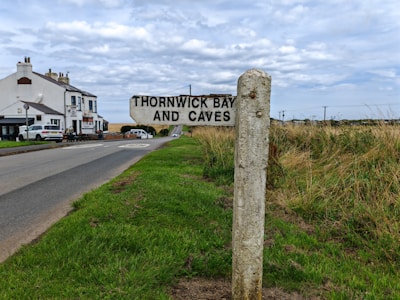 A countryside road with a signpost marking the direction to Thornwick Bay and Caves. The road leads towards a row of white houses, which are surrounded by green fields and dry grass. The sky is overcast with a layer of clouds.