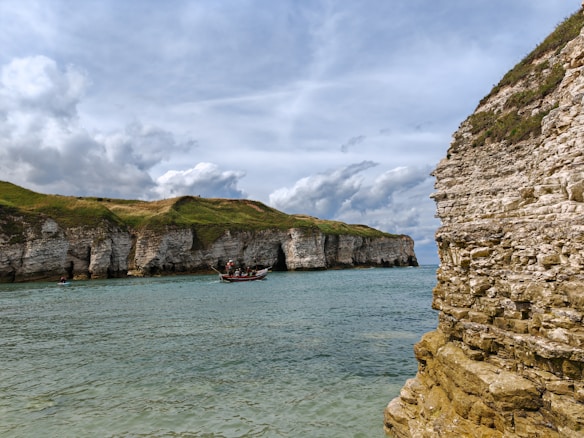 The image features a coastal scene with prominent rocky cliffs and a calm sea beneath a cloudy sky. A small boat with several people on board is in the water near the cliffs. The rocks and grass on the cliffs exhibit earthy tones, creating a natural and tranquil setting.