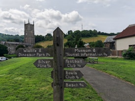 A wooden signpost with various directions is situated in a grassy park. The signs point towards different attractions such as a wildlife park, museum, tourist information, silver mine, football club, bowls club, and a beach, each with distances marked. A church with a tall spire is visible in the background, along with hills, trees, and a partly cloudy sky. Nearby, there are buildings and a couple of benches.