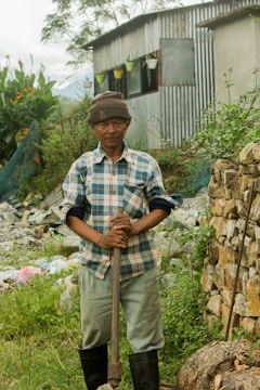 A man wearing a plaid shirt, khaki pants, and a beanie hat stands in a garden holding a gardening tool. Behind him is a corrugated metal shed with potted plants hanging near the window. The surrounding area is lush with greenery, flowers, and a stone wall.