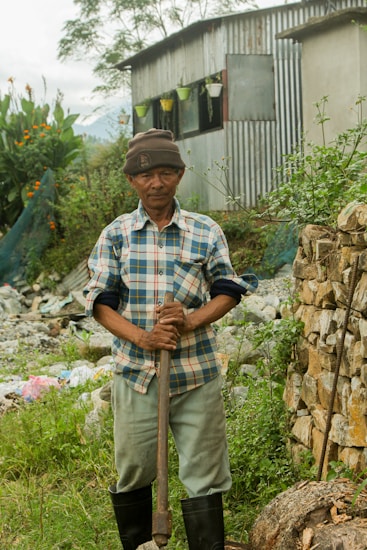 A man wearing a plaid shirt, khaki pants, and a beanie hat stands in a garden holding a gardening tool. Behind him is a corrugated metal shed with potted plants hanging near the window. The surrounding area is lush with greenery, flowers, and a stone wall.