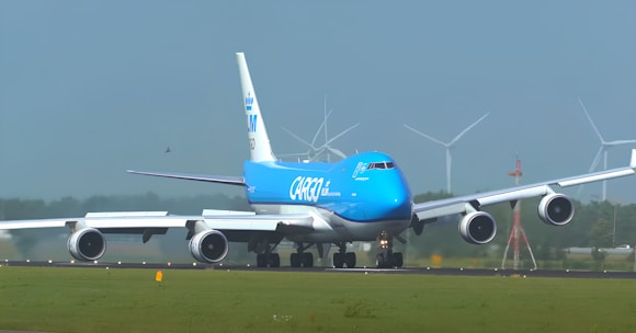 A large cargo airplane is taxiing on an airport runway. It features a bright blue and white color scheme and the words 'KLM Cargo' on its fuselage. The scene includes several wind turbines in the background against a clear blue sky, suggesting a modern and environmentally conscious setting.