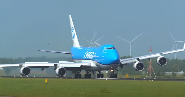 A large cargo airplane is taxiing on an airport runway. It features a bright blue and white color scheme and the words 'KLM Cargo' on its fuselage. The scene includes several wind turbines in the background against a clear blue sky, suggesting a modern and environmentally conscious setting.