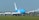 A large cargo airplane is taxiing on an airport runway. It features a bright blue and white color scheme and the words 'KLM Cargo' on its fuselage. The scene includes several wind turbines in the background against a clear blue sky, suggesting a modern and environmentally conscious setting.