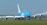 A large cargo airplane is taxiing on an airport runway. It features a bright blue and white color scheme and the words 'KLM Cargo' on its fuselage. The scene includes several wind turbines in the background against a clear blue sky, suggesting a modern and environmentally conscious setting.