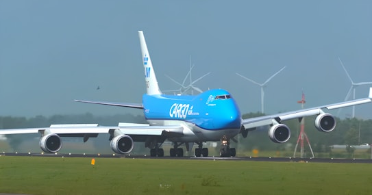 A large cargo airplane is taxiing on an airport runway. It features a bright blue and white color scheme and the words 'KLM Cargo' on its fuselage. The scene includes several wind turbines in the background against a clear blue sky, suggesting a modern and environmentally conscious setting.